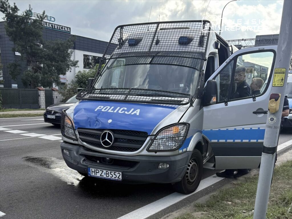 Policyjny furgon z uszkodzoną przednią częścią zatrzymany na ulicy, obok widać przechodnia i sygnalizator.