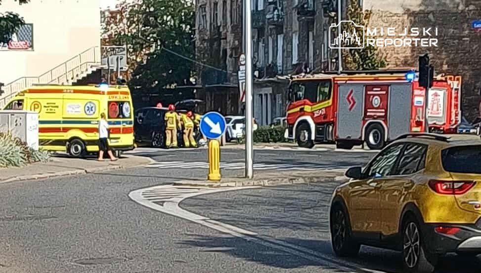 Karetka pogotowia oraz wóz strażacki na skrzyżowaniu, obok grupy ratowników udzielających pomocy.
