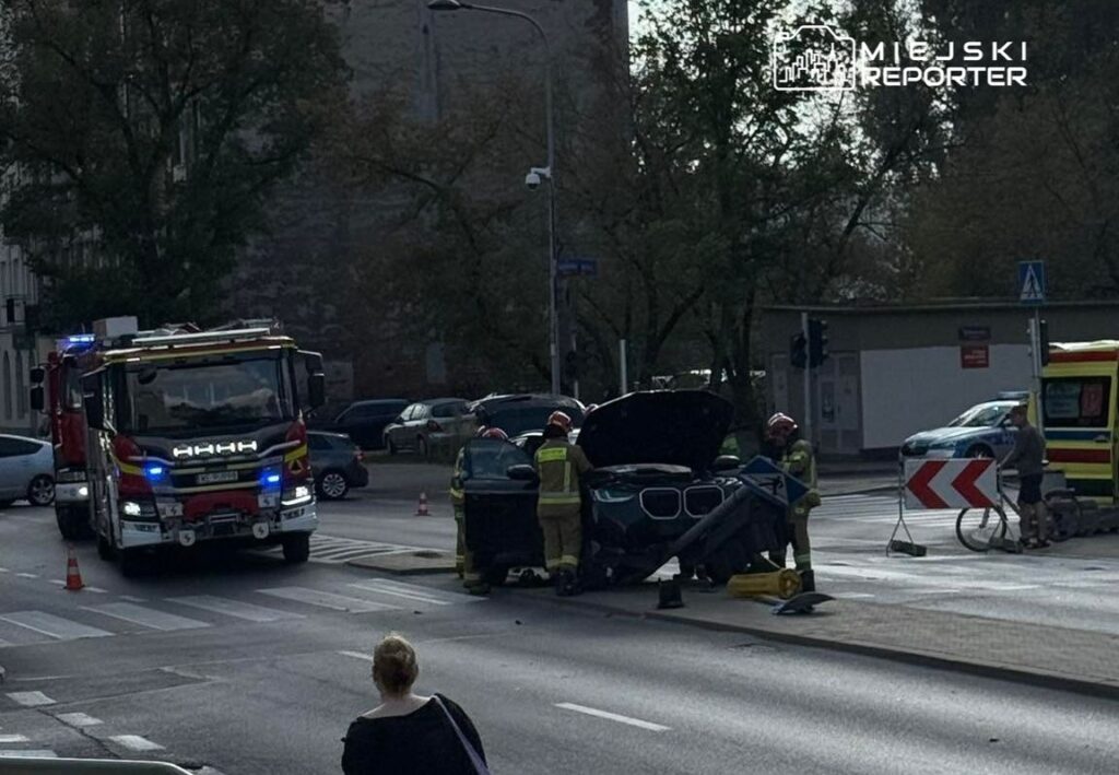 Strażacy pracują nad usunięciem uszkodzonego samochodu na skrzyżowaniu ulicy, obok stojącego wozu strażackiego.