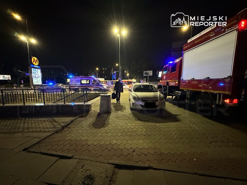 Mężczyzna z torbą idzie w kierunku karetki i wozu strażackiego na oświetlonym placu w nocy.