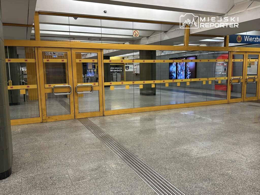 Drzwi stacji metra z przeszkloną fasadą, oznaczone żółtymi taśmami, w tle widać ekran informacyjny.