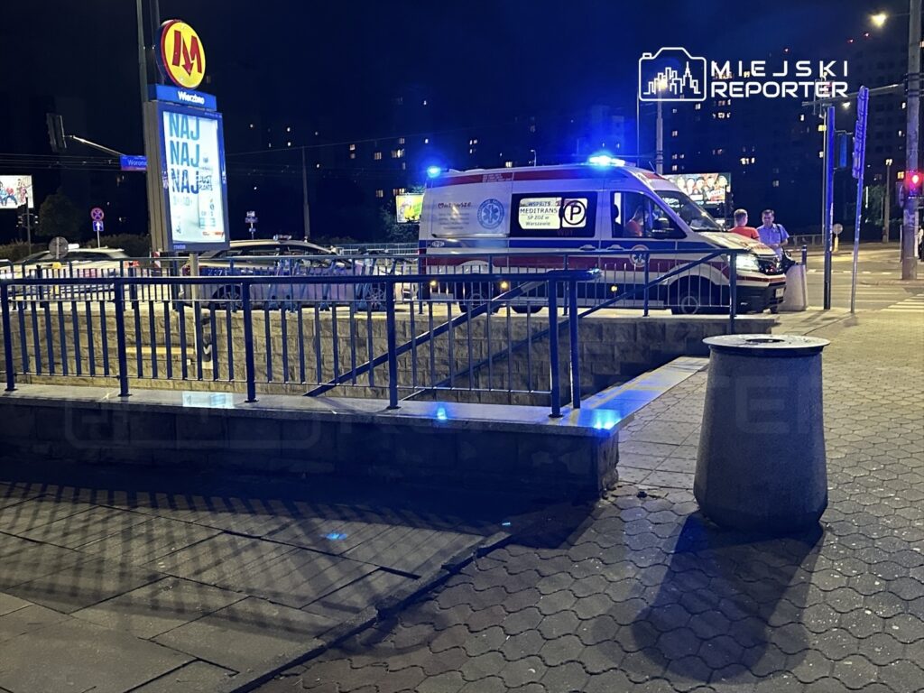 Karetka pogotowia z włączonymi sygnałami świetlnymi stoi przy przejściu dla pieszych w nocy, obok billboardu reklamowego.