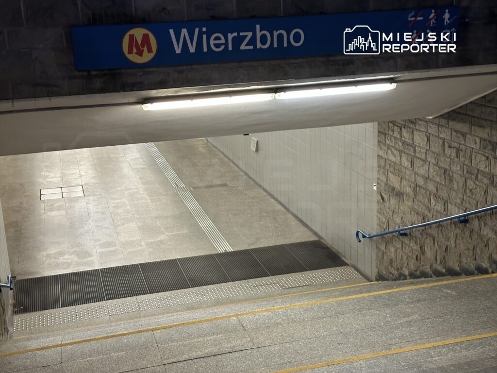 Stacja metra z niebieskim znakiem "Wierzbno", schody prowadzące w dół oraz gładka posadzka.