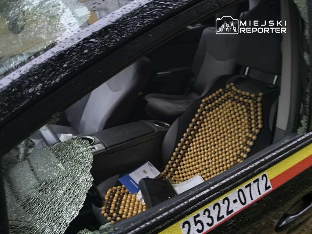 Zniszczona szyba samochodu osobowego, w środku widoczny fotel z drewnianym pokrowcem oraz dokumenty na siedzeniu.