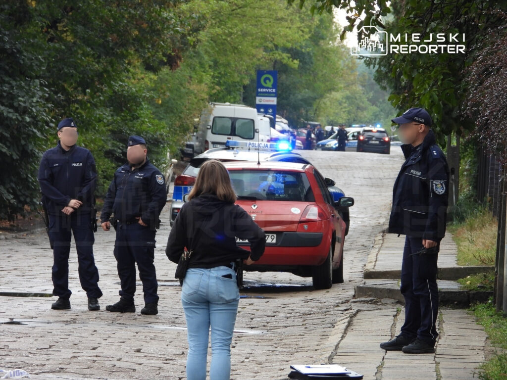 Grupa policjantów w mundurach rozmawia na ulicy, obok zaparkowanego radiowozu, w tle widać inne pojazdy policyjne.