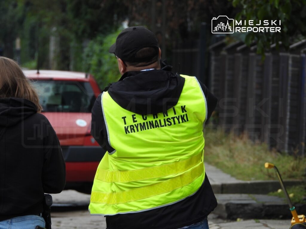 Mężczyzna w kamizelce z napisem "TECHNIK KRYMINALISTYKI" obserwujący miejsce zdarzenia na ulicy.