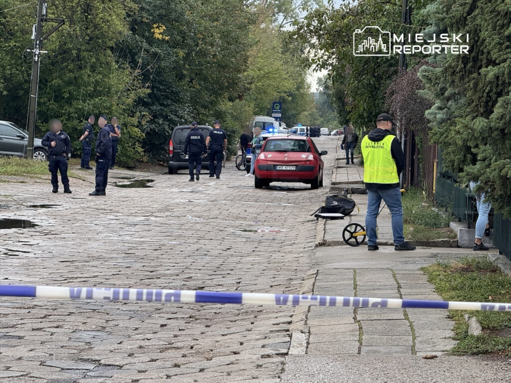 Grupa policjantów prowadzi czynności na ulicy, obok zaparkowanego samochodu i bariery zabezpieczającej.