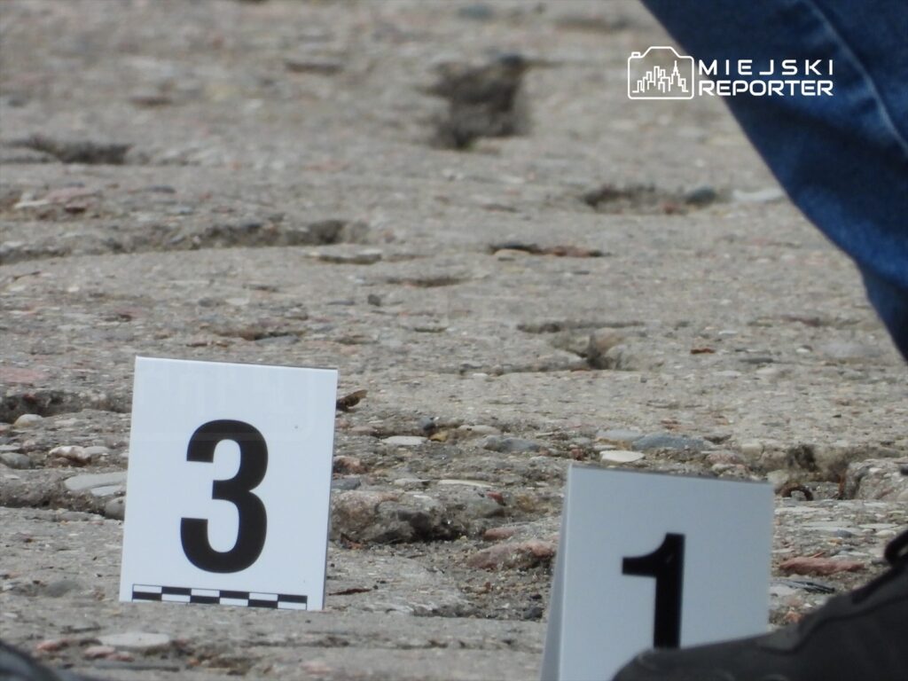 Na nawierzchni ulicy leżą dwa oznaczenia z numerami 1 i 3, obok widoczna stopa w czarnej obuwie.