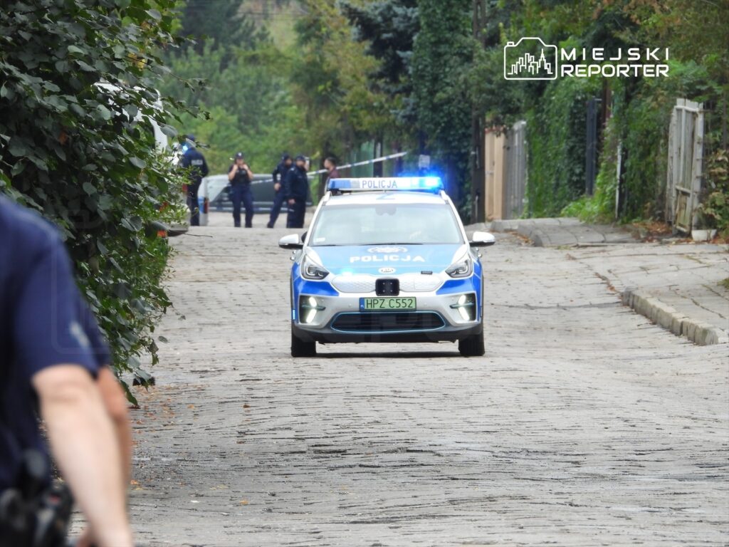 Policyjny radiowóz z włączonymi sygnalizatorami porusza się w wąskiej uliczce, w tle funkcjonariusze w mundurach.