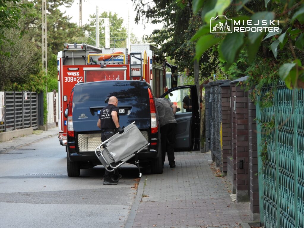 Mężczyzna w mundurze przenosi wózek na chodniku obok zaparkowanego samochodu, w tle widoczna straż pożarna.