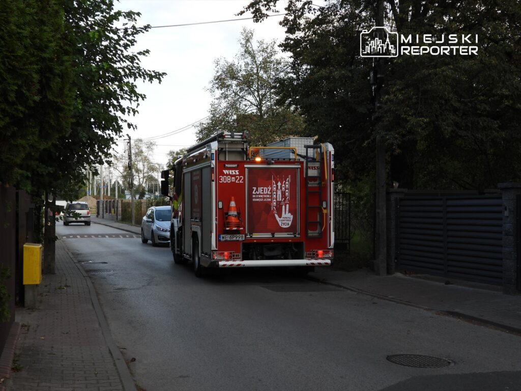 Wóz strażacki wyjeżdża z wąskiej uliczki, obok zaparkowanego samochodu, w otoczeniu drzew.