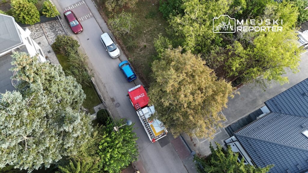 Czerwony wóz strażacki zaparkowany na wąskiej ulicy otoczonej drzewami i budynkami mieszkalnymi.