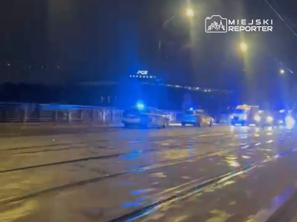 Funkcjonariusze policji oraz karetka pogotowia przejeżdżają po oświetlonym moście w nocy, w tle widoczne niebieskie światła.