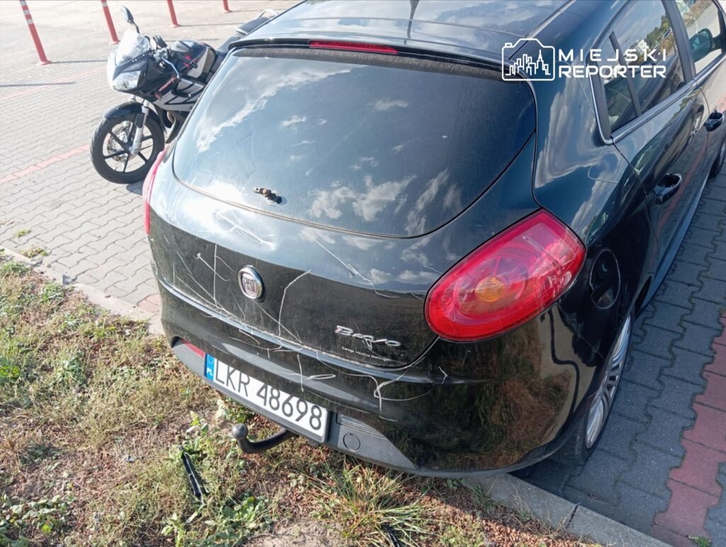Czarny samochód osobowy z widocznymi zadrapaniami zaparkowany obok motocykla na utwardzonej nawierzchni.
