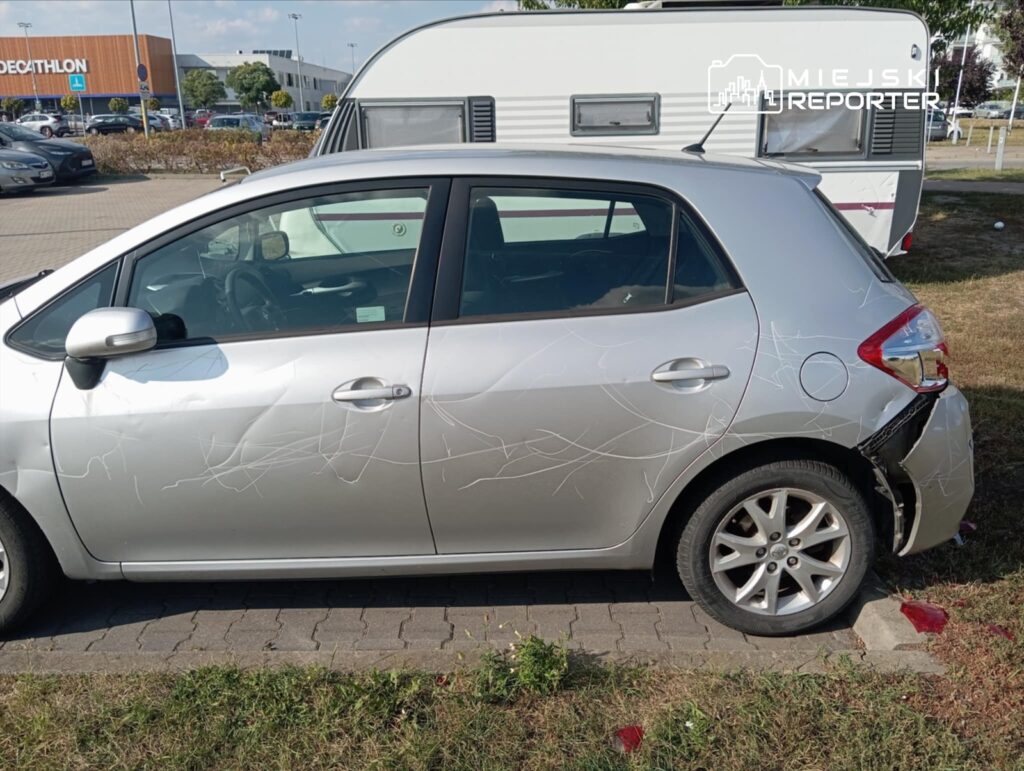 Uszkodzony srebrny samochód osobowy zaparkowany na parkingu, z widocznymi rysami na drzwiach i tylnym zderzaku.