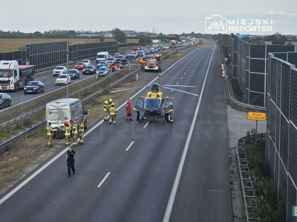 Ratownicy medyczni oraz strażacy udzielają pomocy poszkodowanym przy wypadku drogowym obok lądowiska helikoptera.