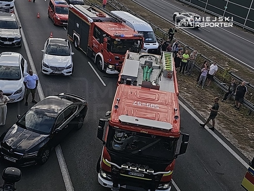 Na jezdni zatrzymane samochody osobowe, obok wóz strażacki z otwartą przestrzenią ładunkową, a w tle grupa ludzi obserwujących sytuację.