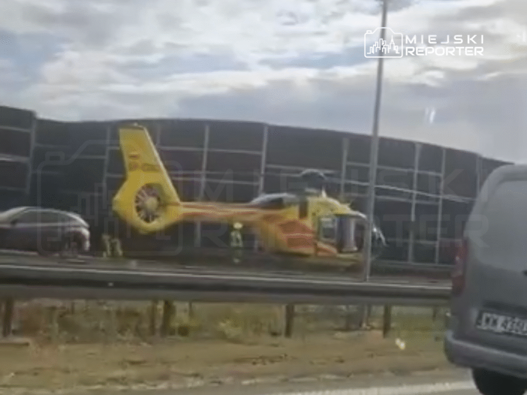 Żółty helikopter lądował na pasie drogowym obok samochodów, a obok niego stoją ratownicy.