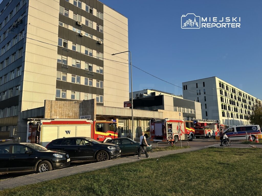 Wielu strażaków i pojazdów ratunkowych przed nowoczesnym budynkiem, w tle niebo o zachodzącym słońcu.