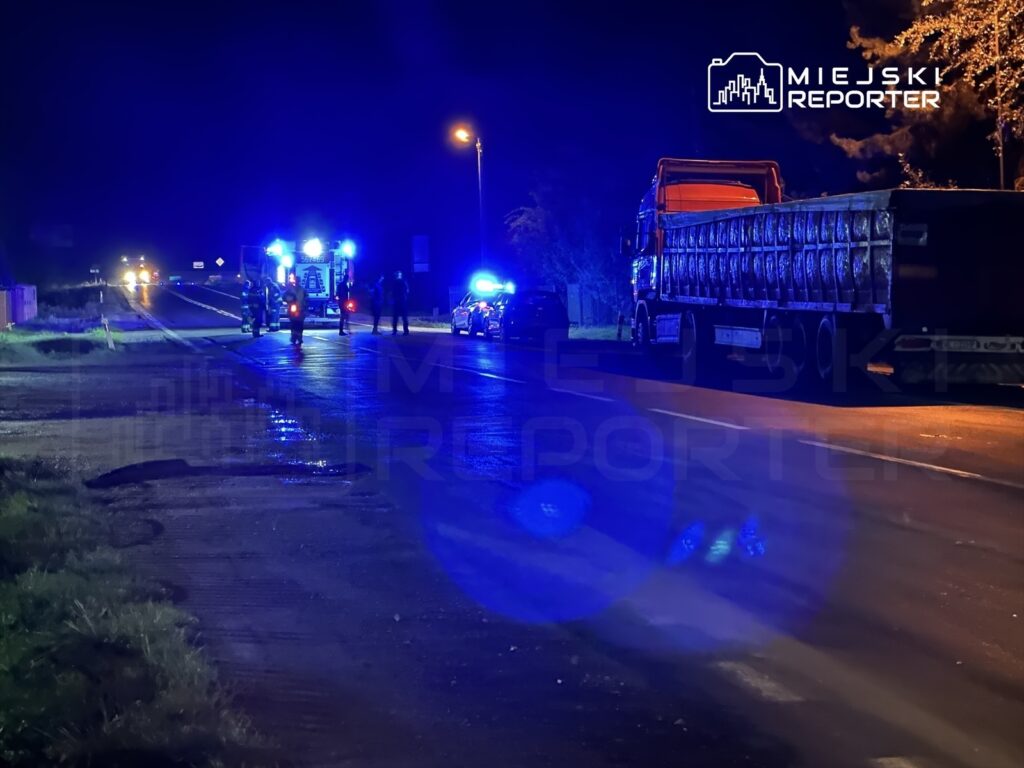 Grupa strażaków oraz policjantów pracuje na drodze w nocy, obok ciężarówki z ładunkiem, w otoczeniu niebieskich świateł.