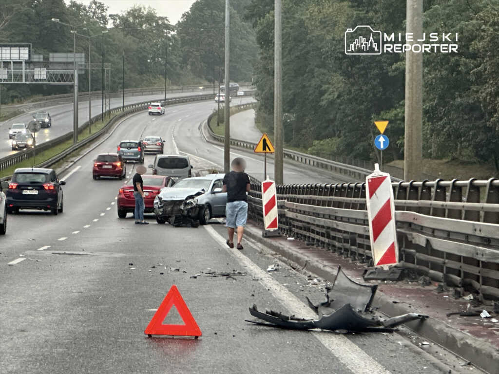 Dwa samochody po kolizji na drodze, mężczyźni sprawdzają uszkodzenia, na jezdni leży rozbita część pojazdu i trójkąt ostrzegawczy.