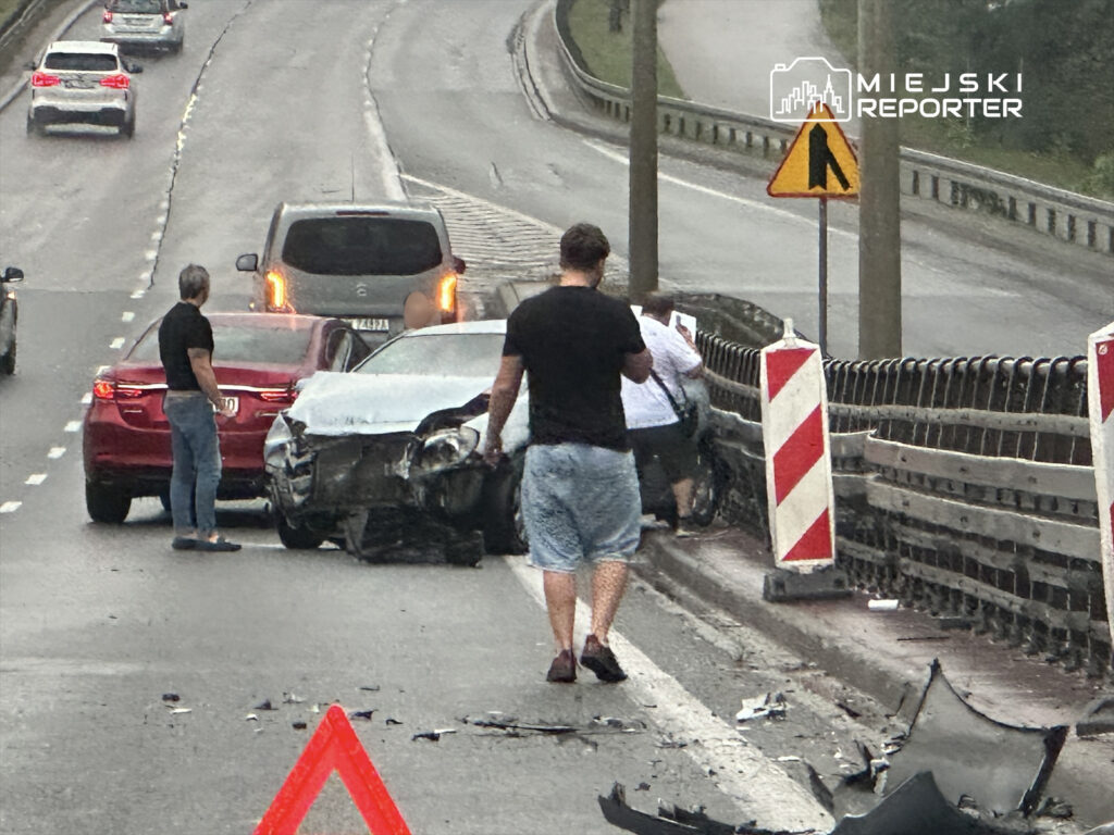Kierowcy zatrzymani obok uszkodzonego samochodu na drodze, obok widoczne znaki drogowe i fragmenty wraku.