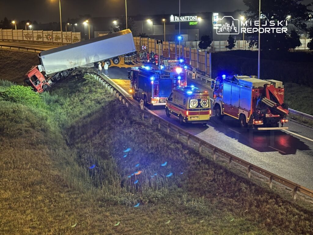 Czerwony ciężarowy pojazd w niebezpiecznej pozycji na poboczu, otoczony przez służby ratunkowe i straż pożarną w nocy.