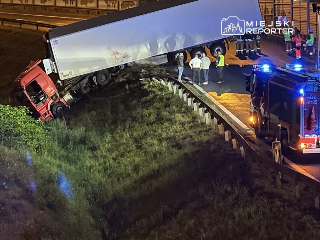 Czerwony ciężarowy pojazd wylegający na pobocze, otoczony przez strażaków i świadków zdarzenia w nocy.