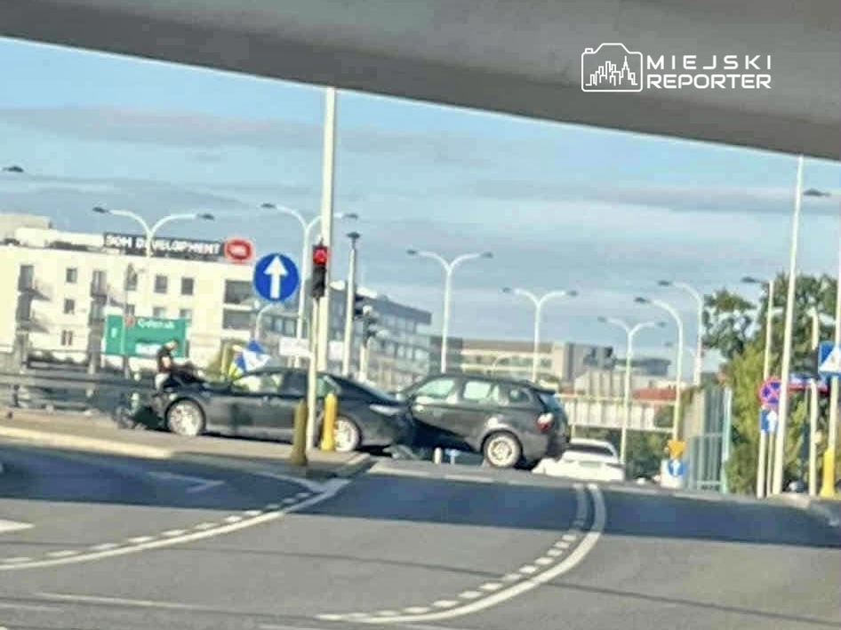 Dwa uszkodzone samochody stoją obok siebie na skrzyżowaniu, a kierowca jednego z nich rozmawia z osobą na zewnątrz.