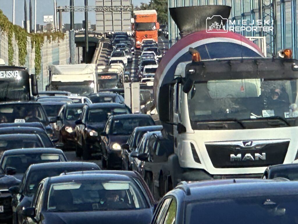 Gęsty ruch samochodowy na drodze, z wieloma pojazdami osobowymi i ciężarówką w centrum.
