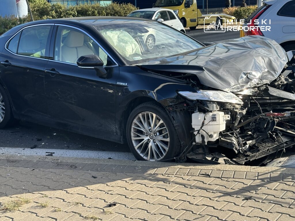 Czarny samochód osobowy z uszkodzoną przednią częścią stoi na poboczu drogi, w tle inne pojazdy.