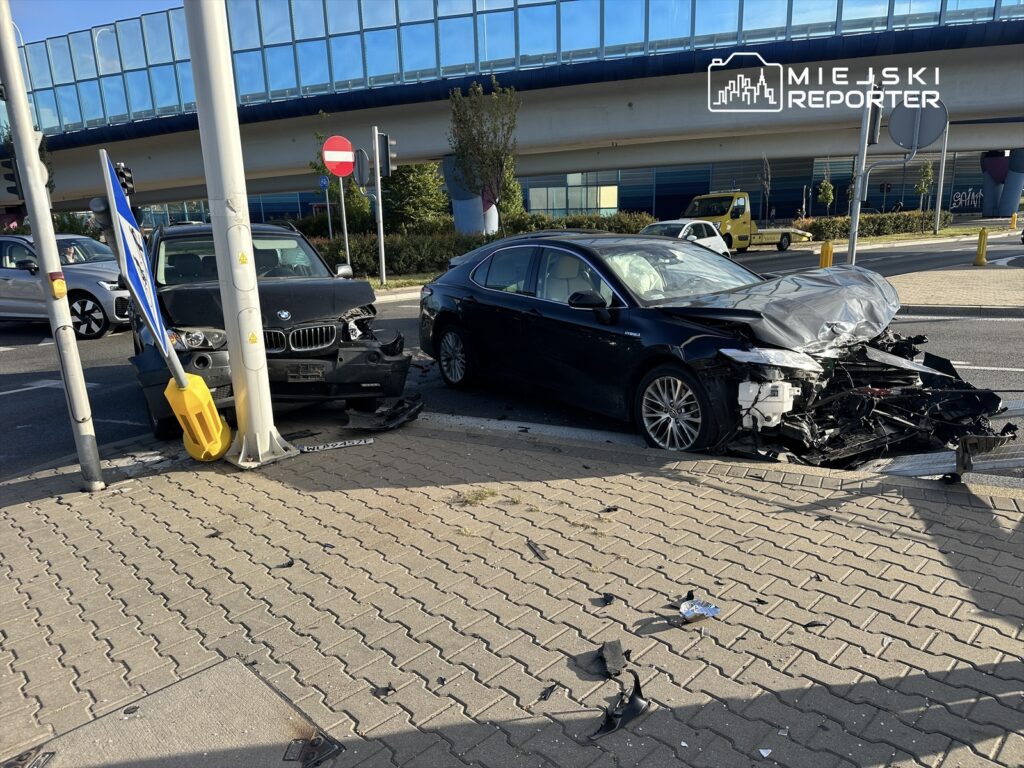 Dwa uszkodzone samochody osobowe zderzone na skrzyżowaniu, jeden z nich z rozbitym przodem, w tle sygnalizacja świetlna.