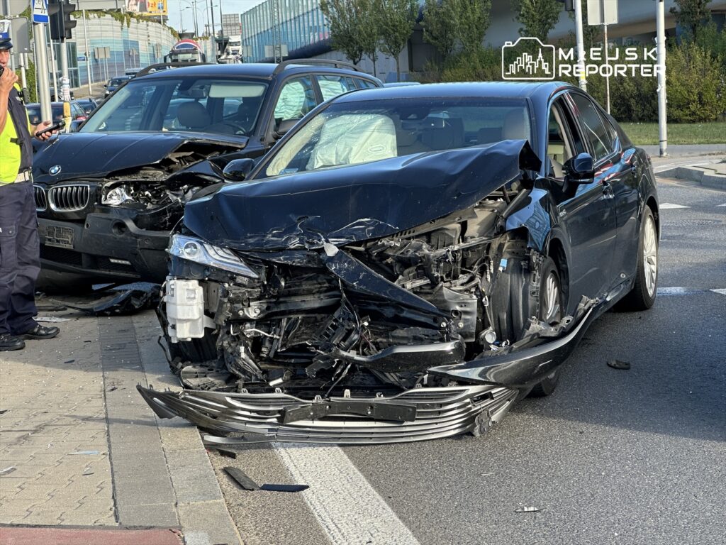 Policyjny funkcjonariusz rozmawia przez telefon obok zderzonych samochodów, jeden z nich ma poważnie uszkodzoną przednią część.