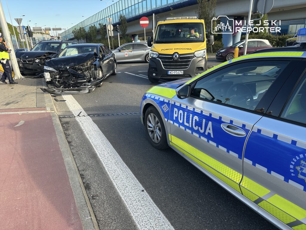 Zderzenie dwóch samochodów osobowych na ulicy, obok policyjnego radiowozu i pojazdu pomocy drogowej.
