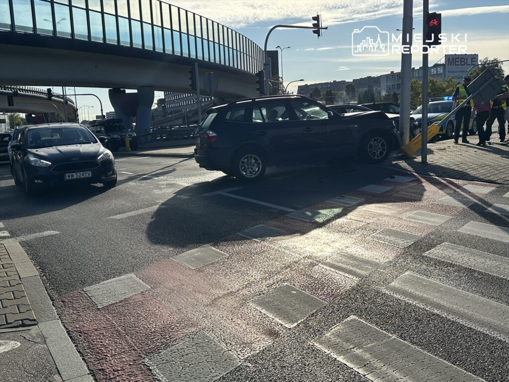 Czarny samochód osobowy zderzony z słupem drogowym na skrzyżowaniu, obok stoją inne pojazdy i przechodnie.