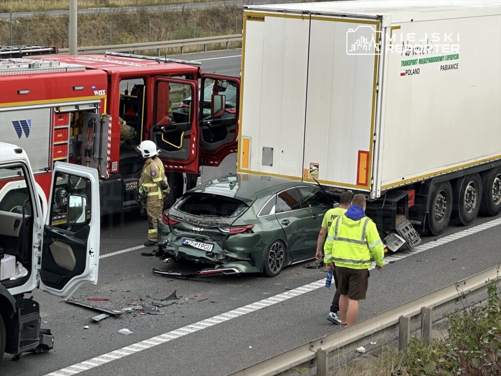 Na drodze stoi czerwona strażacka ciężarówka, obok niej strażak w hełmie, a obok leży rozbity zielony samochód osobowy.