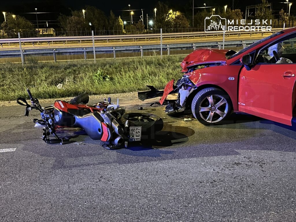 Motocykl leży na jezdni obok uszkodzonego czerwonego samochodu po wypadku drogowym w nocy.