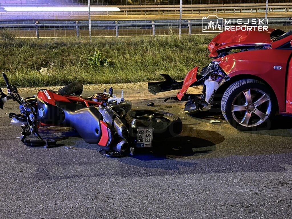Motocykl leżący na drodze obok uszkodzonego czerwonego samochodu po wypadku, w tle pas zieleni.