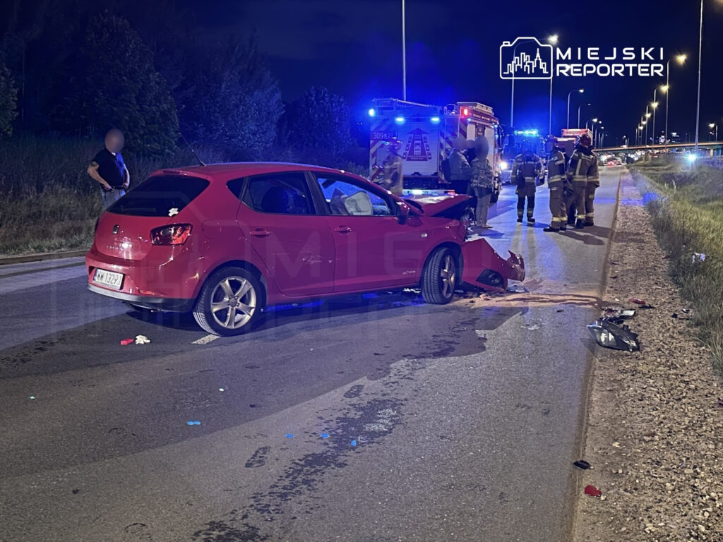 Czerwony samochód osobowy stoi na jezdni po kolizji, wokół znajdują się strażacy i policjanci, a w tle widać niebieskie światła.