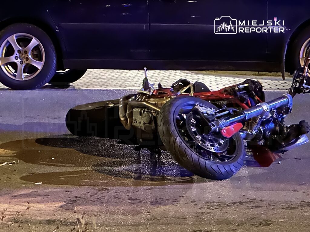 Motocykl leżący na boku na jezdni, obok stoi czarny samochód. Na asfalcie widoczne plamy oleju.