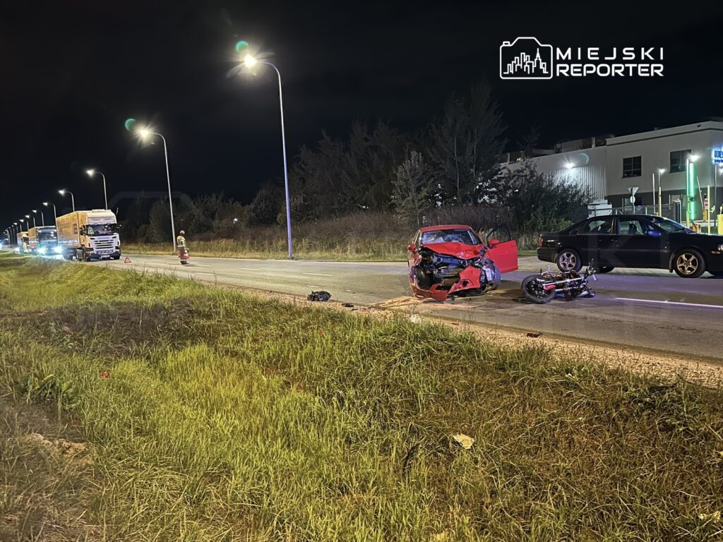 Czerwony samochód osobowy z uszkodzoną przednią częścią stoi na poboczu drogi, obok leży motocykl.