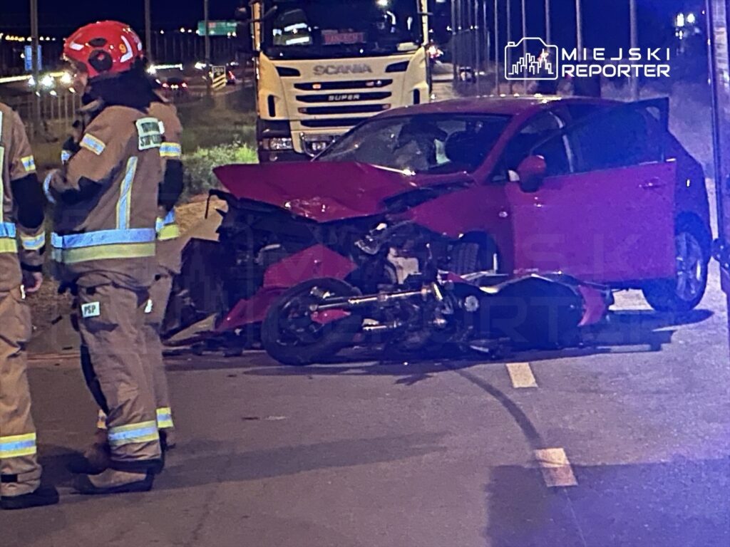 Strażacy w mundurach rozmawiają obok uszkodzonego czerwonego samochodu i przewróconego motocykla na drodze.