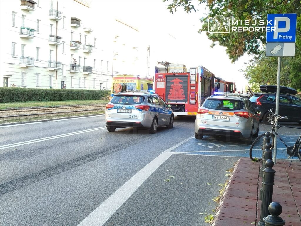 W tle widać nowoczesny budynek mieszkalny, a na pierwszym planie policja i straż pożarna interweniują na jezdni.