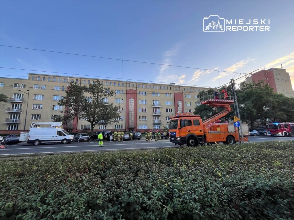 Grupa strażaków w odzieży ochronnej pracuje przy podnośniku w pobliżu bloku mieszkalnego.