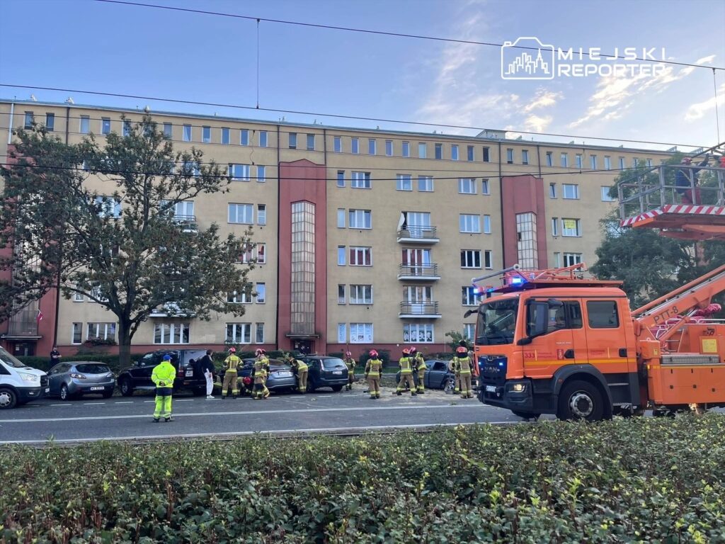 Ratownicy w sprzęcie ochronnym zabezpieczają miejsce zdarzenia przed budynkiem mieszkalnym, obok stoją pojazdy.