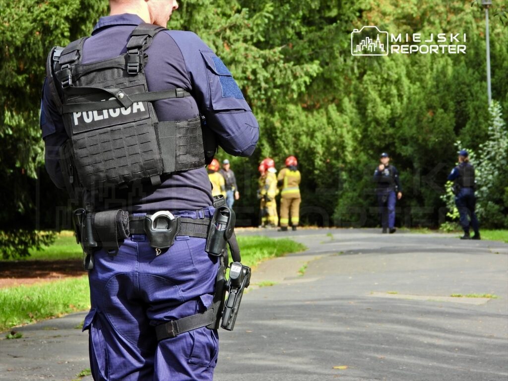 Policjant w kamizelce kuloodpornej obserwuje grupę strażaków i policjantów w parku.