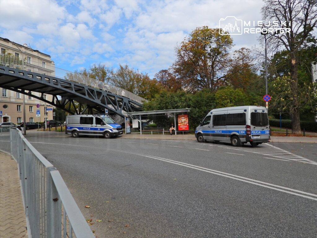 Dwa radiowozy policyjne zatrzymane przy przystanku autobusowym, w tle zadaszony chodnik i drzewa.