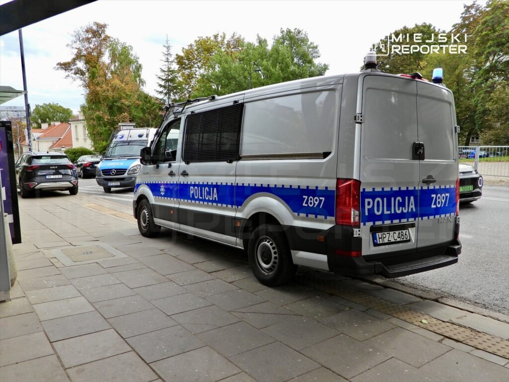 Policyjny pojazd zaparkowany na chodniku, obok innych samochodów, w miejskim otoczeniu z drzewami w tle.