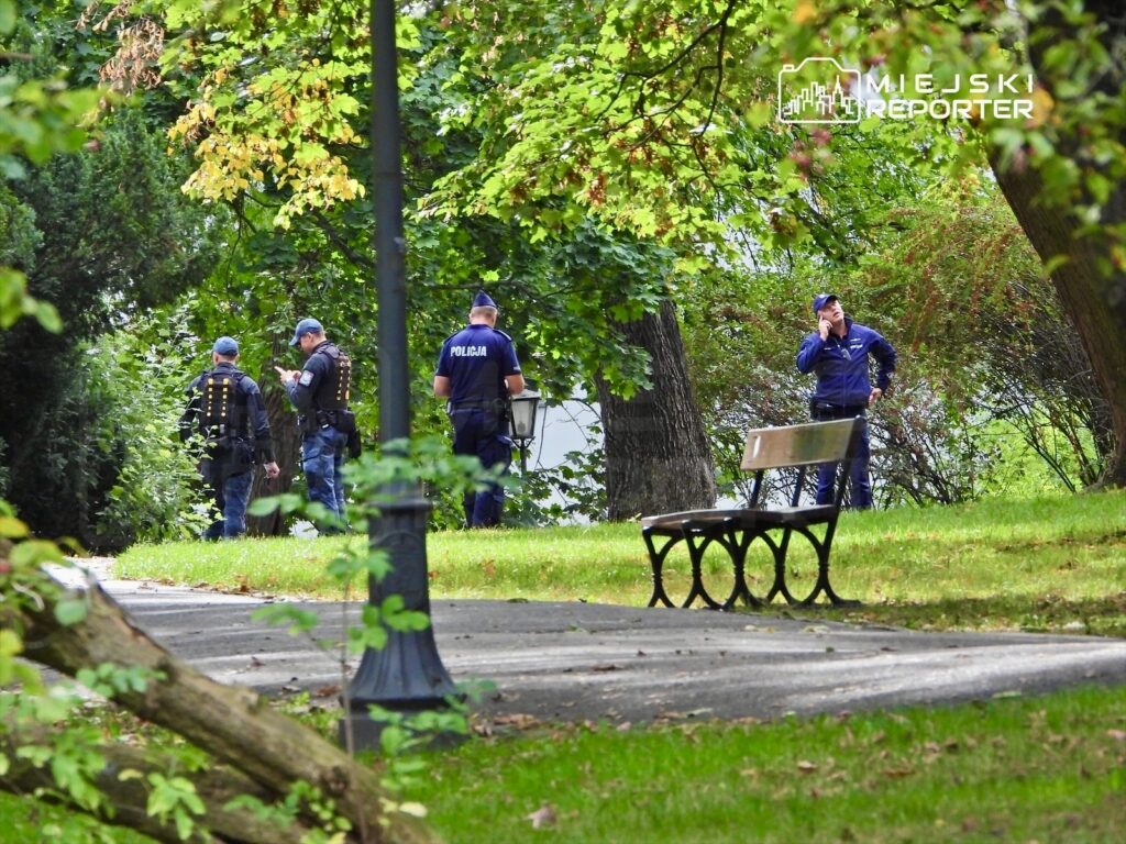 Grupa policjantów w mundurach rozmawia w parku, otoczona zielenią drzew i krzewów.