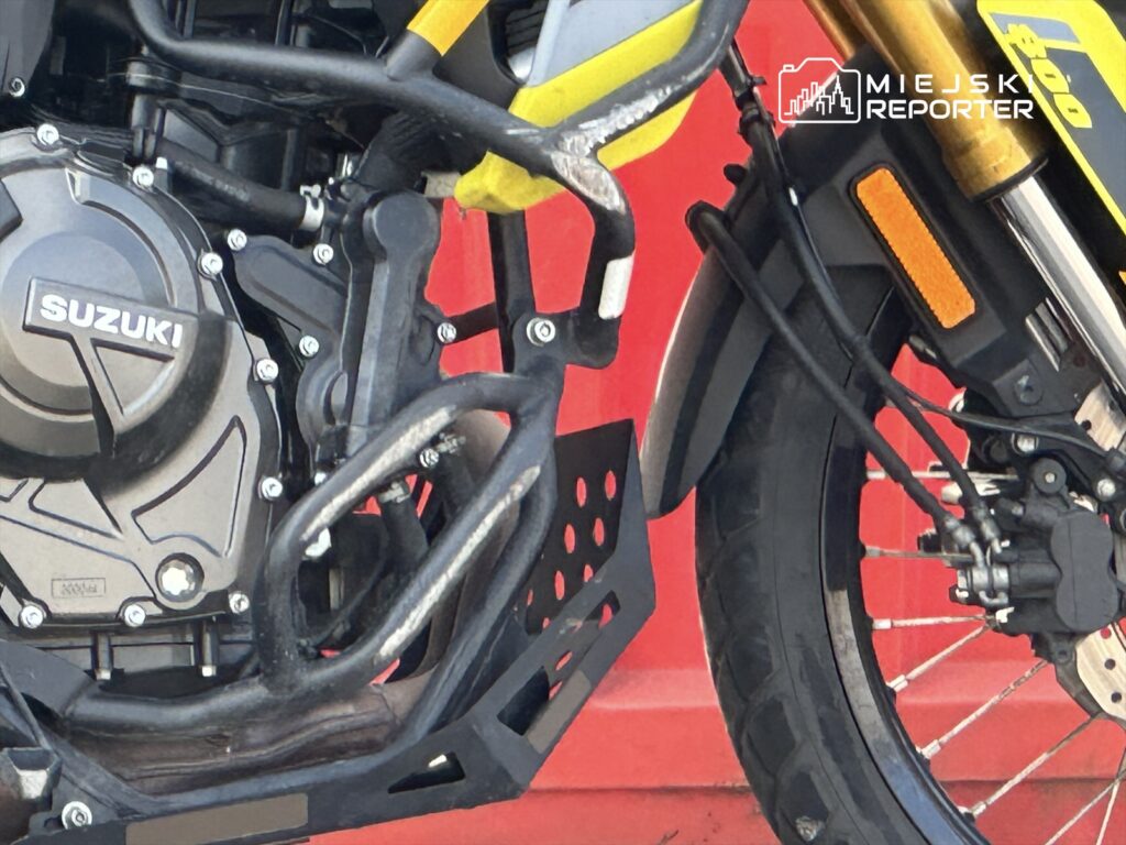 Czarna motocyklowa rama z silnikiem Suzuki, z widocznymi elementami konstrukcyjnymi i oponą, na tle czerwonej ściany.